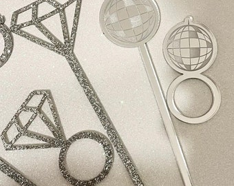 May include: Silver glitter cake toppers featuring a diamond ring and a disco ball. The toppers are attached to sticks.