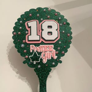 May include: A green glitter cake topper with the number 18 in white with a pink outline. The words "PrettiEE Girl" are below the number. The topper is decorated with small silver and pink beads.