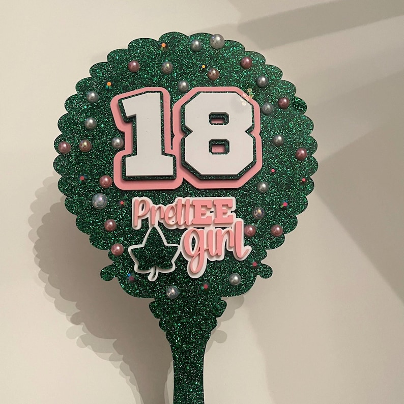 May include: A green glitter cake topper with the number 18 in white with a pink outline. The words "PrettiEE Girl" are below the number. The topper is decorated with small silver and pink beads.