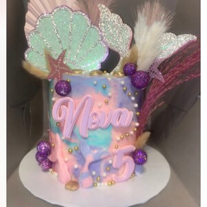 May include: A pastel pink and purple cake decorated with a mermaid tail, seashells, and the name "Nova" in pink letters. The cake is topped with gold and white sprinkles and purple and gold beads.