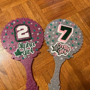 May include: Two glittery hand mirrors. One is magenta with the number 2 and the words "Alpha girl". The other is silver with the number 7 and the words "Pretty girl". Both mirrors have pearl and gem accents.