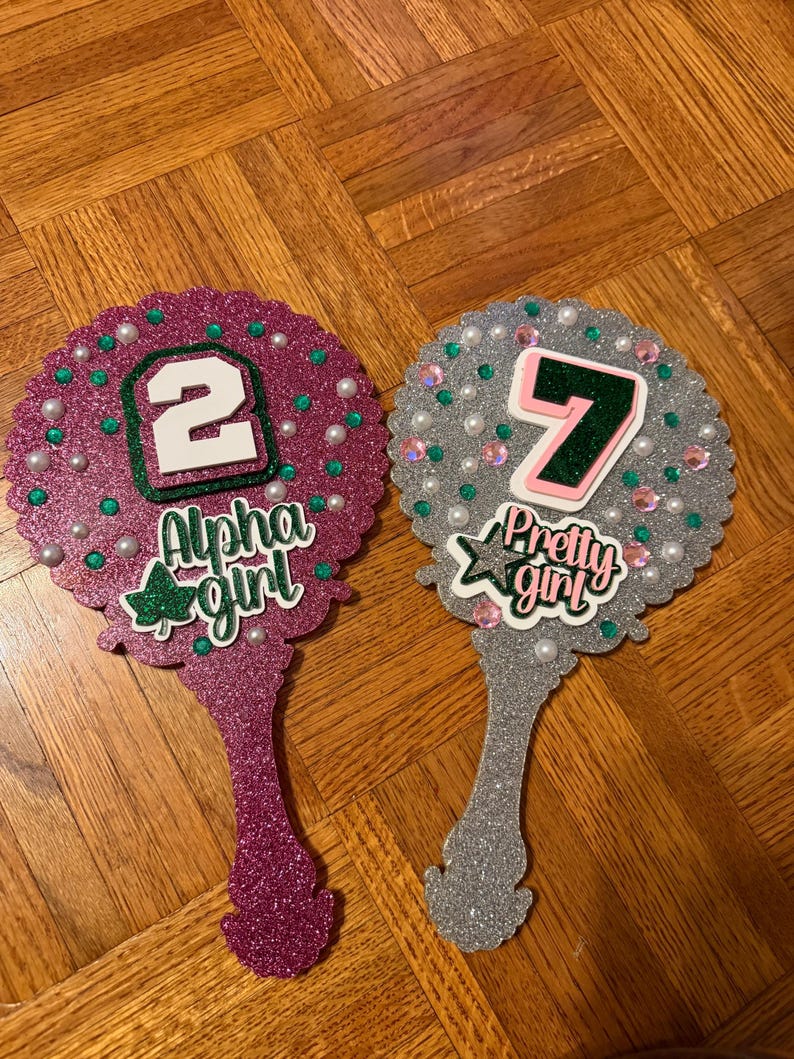 May include: Two glittery hand mirrors. One is magenta with the number 2 and the words "Alpha girl". The other is silver with the number 7 and the words "Pretty girl". Both mirrors have pearl and gem accents.