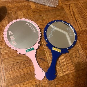 May include: Two hand mirrors, one pink and one blue, with scalloped edges and pearl accents. The pink mirror has the name "Jasmine" and "1908" printed on it. The blue mirror has the name "Brooke" and "1922" printed on it.
