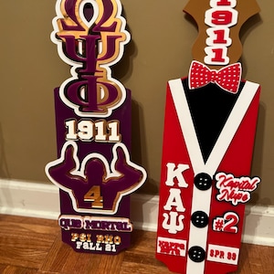 May include: Two decorative wooden plaques. One is purple with gold and white accents, featuring a Greek symbol, the number 1911, and other text. The other is red with black and white accents, with a bow tie and the number 1911.