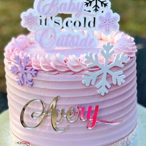 May include: A pink frosted cake with a white snowflake design and a topper that reads "Baby, it's cold outside". The cake is decorated with a silver nameplate that reads "Avery".