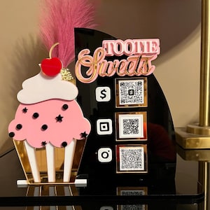May include: A decorative sign for a sweets business, featuring a pink cupcake with a cherry on top. The sign has the text "Tootie Sweets" in pink and gold, along with QR codes and social media icons on a black background.