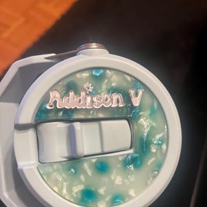 May include: A light blue water bottle with a white and blue speckled lid. The lid has a slide switch and a silver sticker with the name "Addison V" on it.