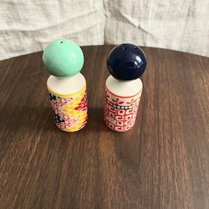 May include: A pair of decorative salt and pepper shakers. One shaker has a light green ball top, the other has a dark blue ball top. The bases are cylindrical with colorful geometric and floral patterns. The shakers are on a dark wood surface.