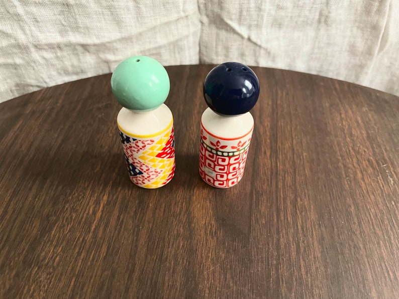 May include: A pair of decorative salt and pepper shakers. One shaker has a light green ball top, the other has a dark blue ball top. The bases are cylindrical with colorful geometric and floral patterns. The shakers are on a dark wood surface.