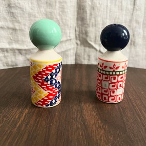 May include: Two decorative ceramic salt and pepper shakers. One has a mint green ball top and a patterned body with red, yellow, and blue designs. The other has a navy blue ball top and a red and white geometric pattern.
