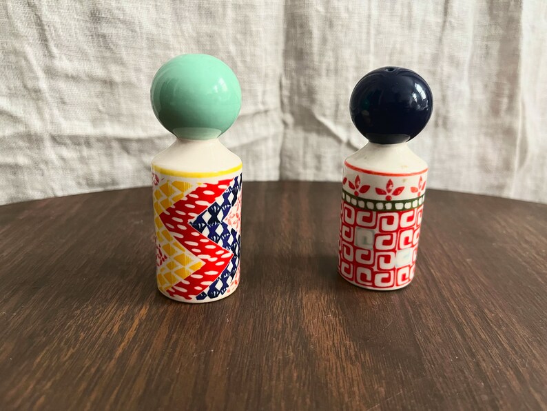 May include: Two decorative ceramic salt and pepper shakers. One has a mint green ball top and a patterned body with red, yellow, and blue designs. The other has a navy blue ball top and a red and white geometric pattern.
