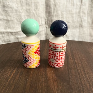 May include: Two decorative salt and pepper shakers. One has a mint green ball top, the other a navy blue ball top. The bodies are white with colorful geometric patterns in red, yellow, and blue. They sit on a dark wooden surface.