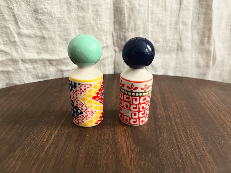 May include: Two decorative salt and pepper shakers. One has a mint green ball top, the other a navy blue ball top. The bodies are white with colorful geometric patterns in red, yellow, and blue. They sit on a dark wooden surface.