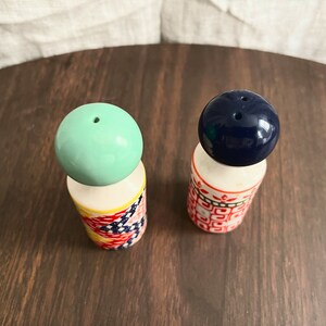 May include: A pair of vintage salt and pepper shakers. One shaker has a light green ball top, the other has a dark blue ball top. The cylindrical bases are decorated with colorful patterns. The shakers are on a dark wooden surface.