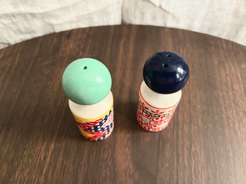 May include: A pair of vintage salt and pepper shakers. One shaker has a light green ball top, the other has a dark blue ball top. The cylindrical bases are decorated with colorful patterns. The shakers are on a dark wooden surface.