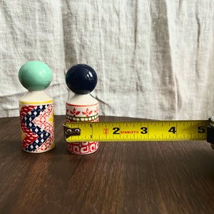 May include: Two decorative ceramic salt and pepper shakers. One shaker has a blue-green ball top, the other has a navy blue ball top. The first shaker has a colorful patchwork design, and the second has a red and green floral pattern. The smaller shaker is approximately 1.5 inches in height.