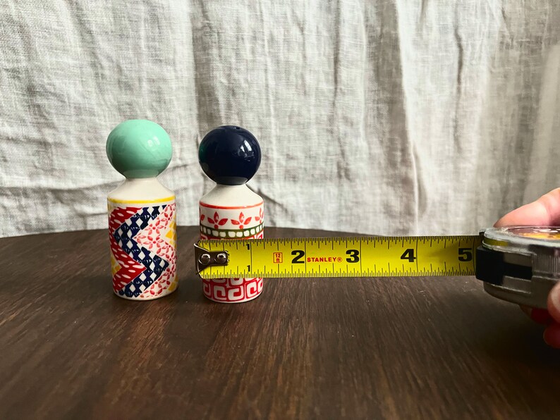 May include: Two decorative ceramic salt and pepper shakers. One shaker has a blue-green ball top, the other has a navy blue ball top. The first shaker has a colorful patchwork design, and the second has a red and green floral pattern. The smaller shaker is approximately 1.5 inches in height.