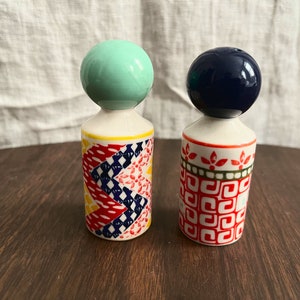 May include: Two decorative ceramic salt and pepper shakers. One has a mint green ball top, the other a navy blue ball top. The bodies are white with colorful geometric patterns in red, yellow, and blue. The shakers are on a dark wooden surface.