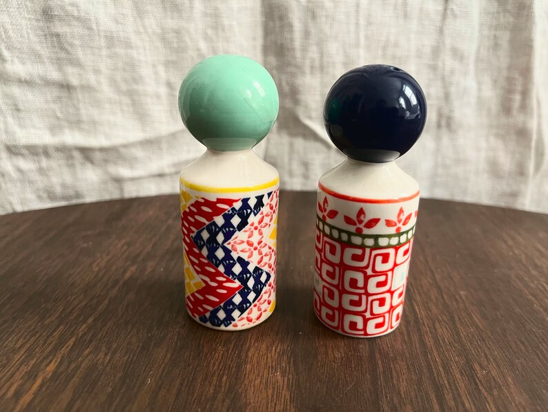 May include: Two decorative ceramic salt and pepper shakers. One has a mint green ball top, the other a navy blue ball top. The bodies are white with colorful geometric patterns in red, yellow, and blue. The shakers are on a dark wooden surface.