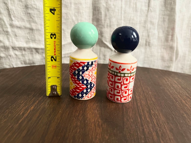 May include: A pair of ceramic salt and pepper shakers. One shaker has a mint green ball top and a colorful geometric pattern. The other has a navy blue ball top and a red and white pattern. Each shaker is approximately 3 inches tall.