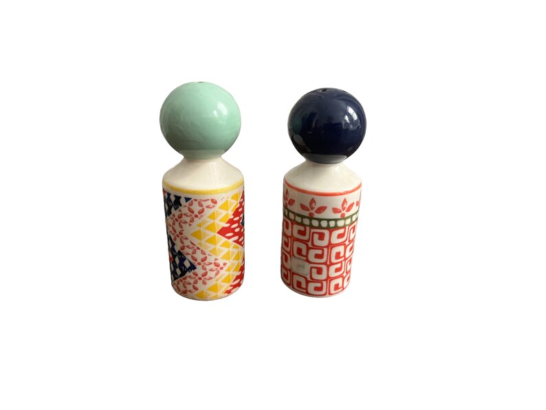 May include: A pair of ceramic salt and pepper shakers. One shaker has a light green ball-shaped top and a body decorated with a colorful geometric pattern. The other shaker has a dark blue ball-shaped top and a body with a red and green pattern.