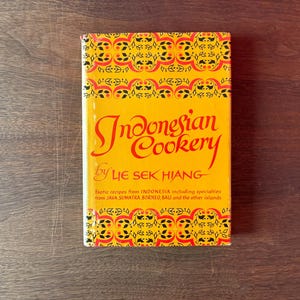 May include: A hardcover cookbook titled "Indonesian Cookery" by Lie Sek Hiang. The book has a yellow cover with red and black decorative patterns. The title is in large, red, cursive script.