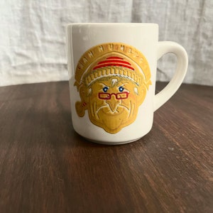 May include: White ceramic mug with a handle, featuring a cartoon image of a man with a hat, glasses, and a pipe. The mug has the words "WAH HUM DUG" in a semi-circle above the man's head.