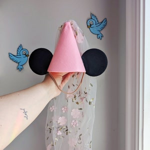 May include: Pink felt party hat with black felt Mickey Mouse ears and a sheer veil with pink floral embroidery.