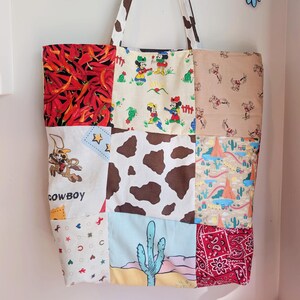 May include: A patchwork tote bag with a variety of fabric patterns, including red chili peppers, Mickey Mouse, cowboy motifs, cow spots, and a desert scene. The handles are white with brown cow spots. Two blue bird decorations are on the wall.