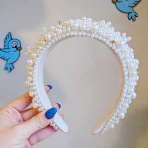 May include: A white headband with a thick layer of white pearls. The headband is slightly curved and has a smooth, satin-like finish.