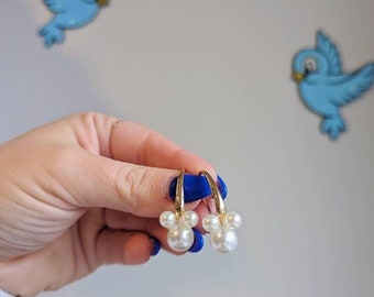 May include: Gold hoop earrings with a cluster of white faux pearls. The earrings are held up against a light gray background with two blue bird decorations.