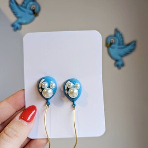 May include: A pair of blue balloon-shaped earrings with white pearl accents and gold-colored wire. The earrings are displayed on a white card, with two blue bird decorations in the background.
