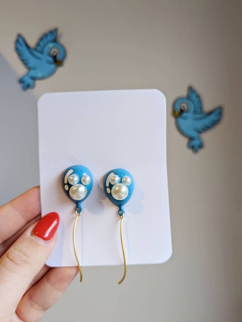 May include: A pair of blue balloon-shaped earrings with white pearl accents and gold-colored wire. The earrings are displayed on a white card, with two blue bird decorations in the background.