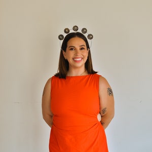 K&ouml;nnte beinhalten: Eine Frau in einem leuchtend orangefarbenen, &auml;rmellosen Kleid mit einem silbernen Stirnband, das mit goldenen runden Verzierungen verziert ist. Sie l&auml;chelt und hat die H&auml;nde auf den H&uuml;ften.