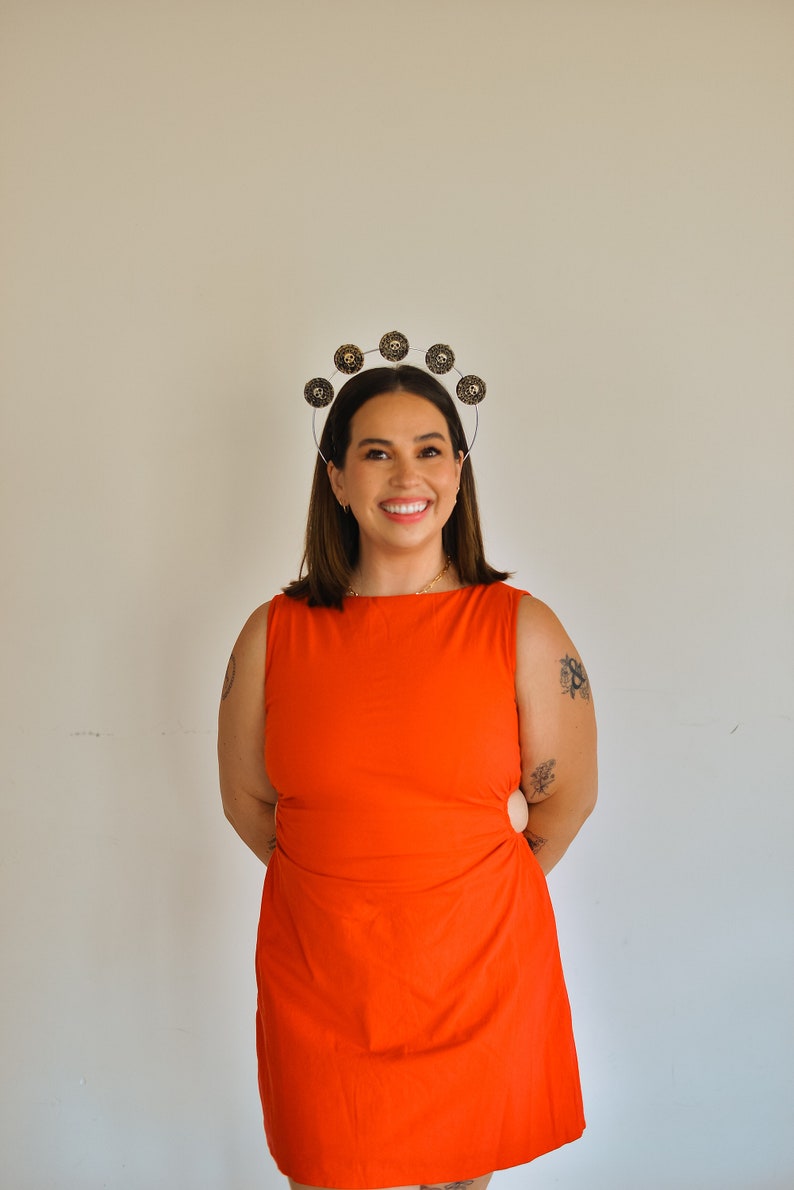 K&ouml;nnte beinhalten: Eine Frau in einem leuchtend orangefarbenen, &auml;rmellosen Kleid mit einem silbernen Stirnband, das mit goldenen runden Verzierungen verziert ist. Sie l&auml;chelt und hat die H&auml;nde auf den H&uuml;ften.
