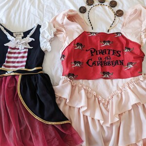 K&ouml;nnte beinhalten: Zwei M&auml;dchenkost&uuml;me, eines ein rot-schwarzes Piratenkost&uuml;m mit einem gestreiften Oberteil und goldenen Verzierungen, das andere ein rosa Kleid mit einem roten Oberteil, auf dem "Pirates of the Caribbean" mit Mickey-Maus-Figuren steht.