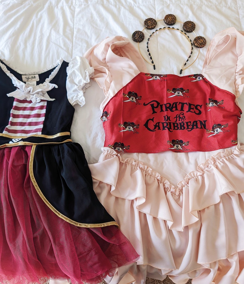K&ouml;nnte beinhalten: Zwei M&auml;dchenkost&uuml;me, eines ein rot-schwarzes Piratenkost&uuml;m mit einem gestreiften Oberteil und goldenen Verzierungen, das andere ein rosa Kleid mit einem roten Oberteil, auf dem "Pirates of the Caribbean" mit Mickey-Maus-Figuren steht.