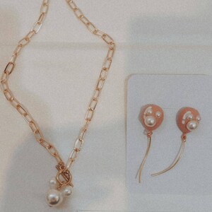 May include: Gold-toned chain necklace with a pearl pendant and matching earrings. The earrings feature peach-colored balloon-shaped charms with pearl accents. The jewelry is displayed on a white surface.