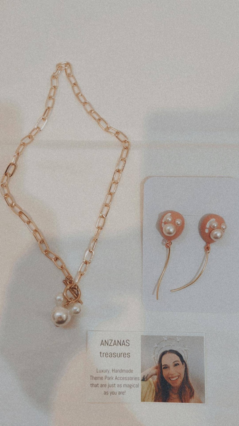 May include: Gold-toned chain necklace with a pearl pendant and matching earrings. The earrings feature peach-colored balloon-shaped charms with pearl accents. The jewelry is displayed on a white surface.