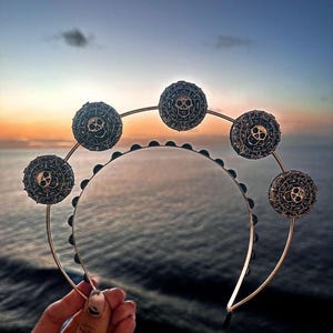 K&ouml;nnte beinhalten: Ein goldenes Stirnband mit schwarzen Totenkopf- und Knochenanh&auml;ngern. Das Stirnband ist mit schwarzen Perlen verziert und hat ein kreisf&ouml;rmiges Design.