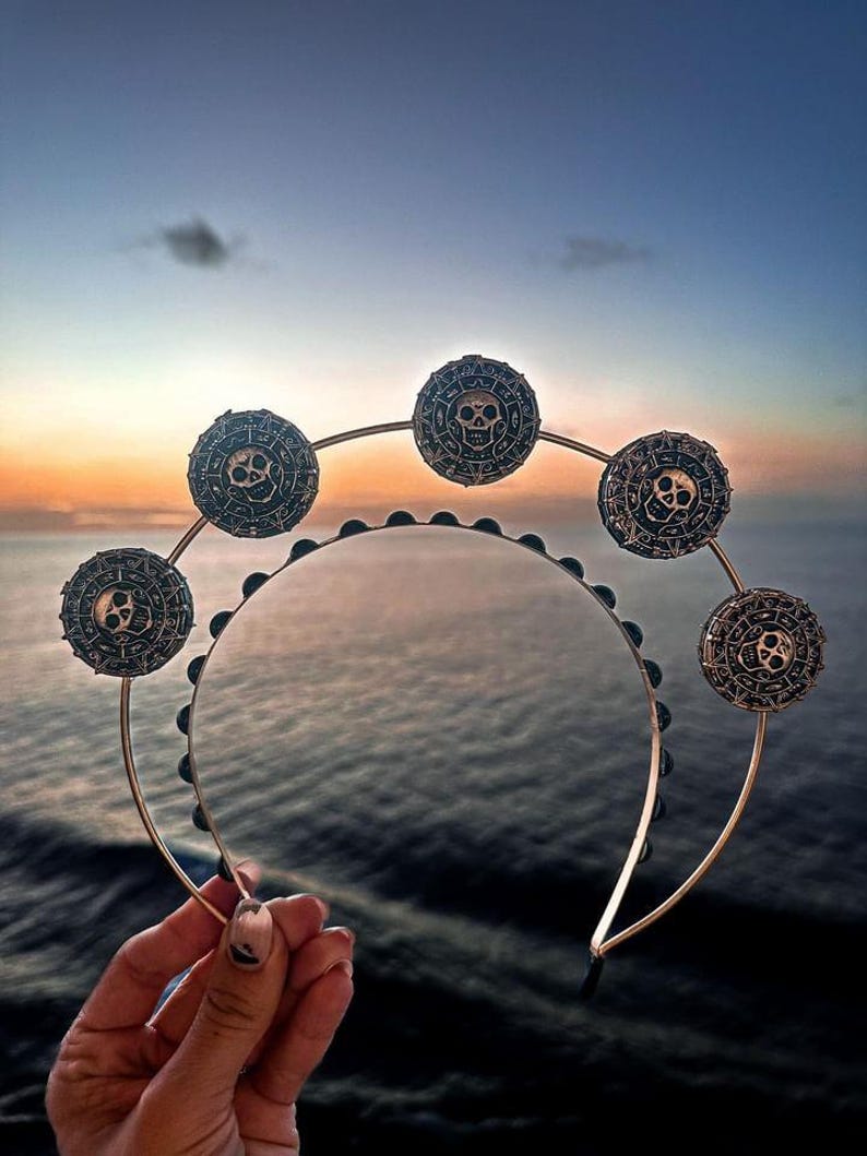 K&ouml;nnte beinhalten: Ein goldenes Stirnband mit schwarzen Totenkopf- und Knochenanh&auml;ngern. Das Stirnband ist mit schwarzen Perlen verziert und hat ein kreisf&ouml;rmiges Design.