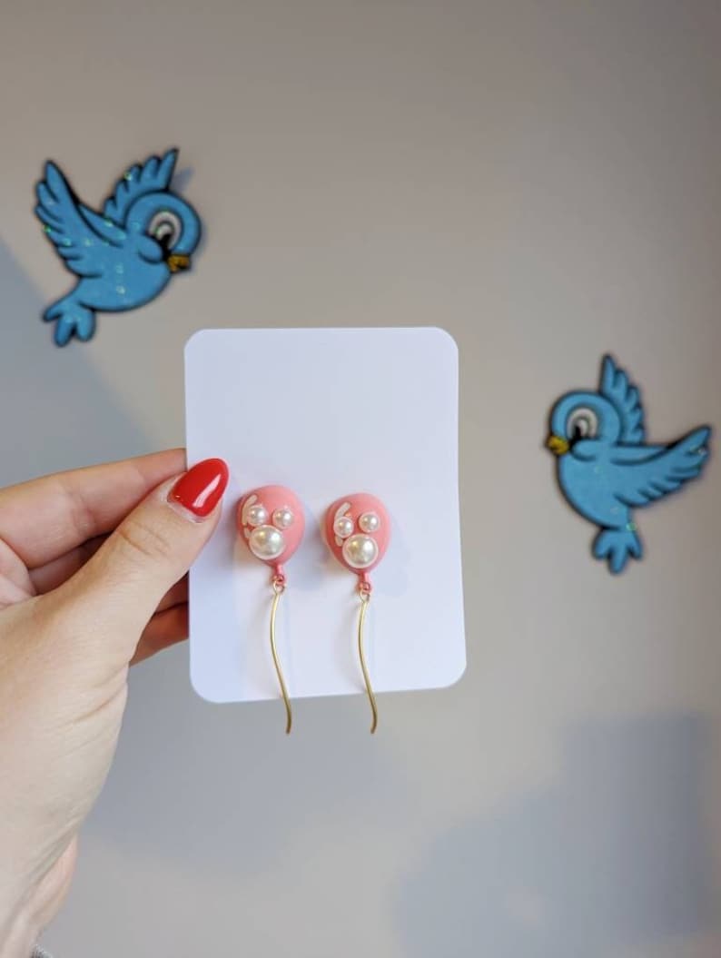 May include: Pink balloon-shaped earrings with white pearl accents and gold-colored curved wires. The earrings are displayed on a white card, with two blue bird decorations in the background.