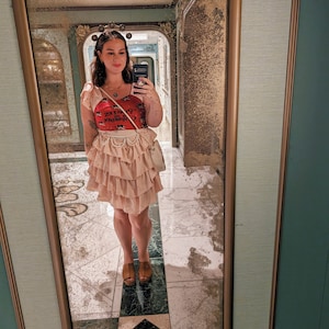 K&ouml;nnte beinhalten: Eine Frau tr&auml;gt einen rosa Faltenrock, ein rotes Tanktop mit einem grafischen Aufdruck, der "Cigarette Queen" sagt, und eine silberne Halskette. Sie steht vor einem Spiegel mit goldenem Rahmen.