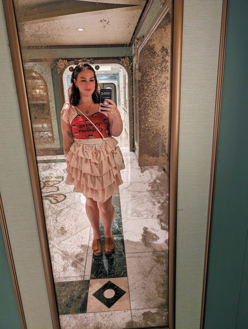 K&ouml;nnte beinhalten: Eine Frau tr&auml;gt einen rosa Faltenrock, ein rotes Tanktop mit einem grafischen Aufdruck, der "Cigarette Queen" sagt, und eine silberne Halskette. Sie steht vor einem Spiegel mit goldenem Rahmen.