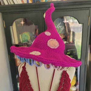 May include: A vibrant magenta crochet mushroom hat with a curved stem and a wide brim. The hat features cream-colored spots and a scalloped edge with blue, green, and yellow accents. The hat is part of a costume.