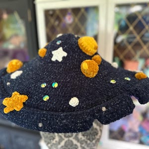 May include: A dark blue, textured crochet hat with white star and circle accents, and yellow, fuzzy, circular accents. Iridescent sequins and small white beads are scattered across the hat. The hat has a wide, wavy brim.