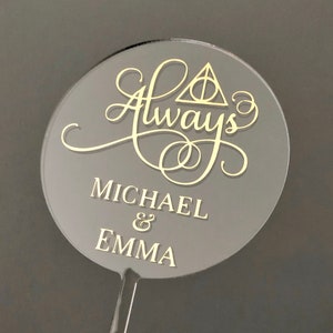 May include: A round, clear acrylic cake topper with the words "Always" in gold cursive script, a gold triangle symbol, and the names "MICHAEL & EMMA" in gold lettering.