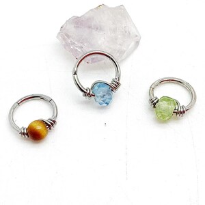 Puede incluir: Tres aros de metal plateado, cada uno con una cuenta de piedra preciosa asegurada con alambre enrollado. Un aro tiene una cuenta de ojo de tigre marrón, otro una piedra azul claro facetada y el tercero una cuenta de piedra bruta verde pálido.