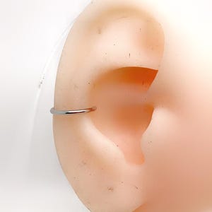 Può includere: Nella foto è presente un orecchino a cerchio argentato. L'orecchino, dal design circolare semplice, è posizionato su un modello di orecchio di colore chiaro. L'orecchino è un piccolo gioiello minimalista.