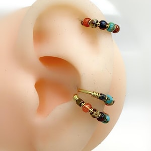 Peut inclure: Deux créoles dorées ornées de petites perles multicolores dans des tons orange, turquoise, marron et bleu foncé. Les perles sont disposées le long du fil de chaque créole, qui est fixée par un détail en fil enroulé au point de fermeture.